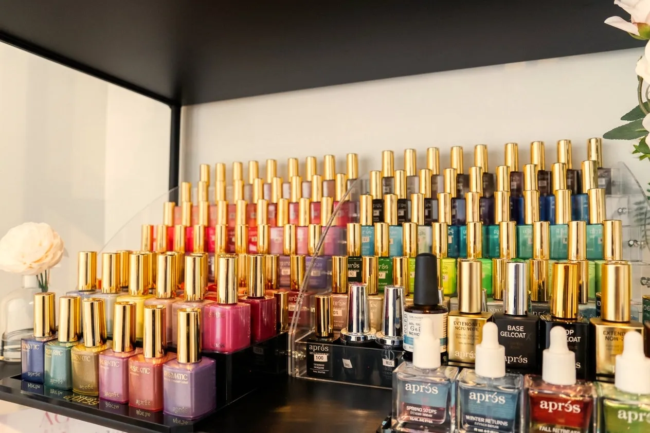 Rows of gel polish bottles with gold caps, arranged by colour on a shelf beside a vase of white roses