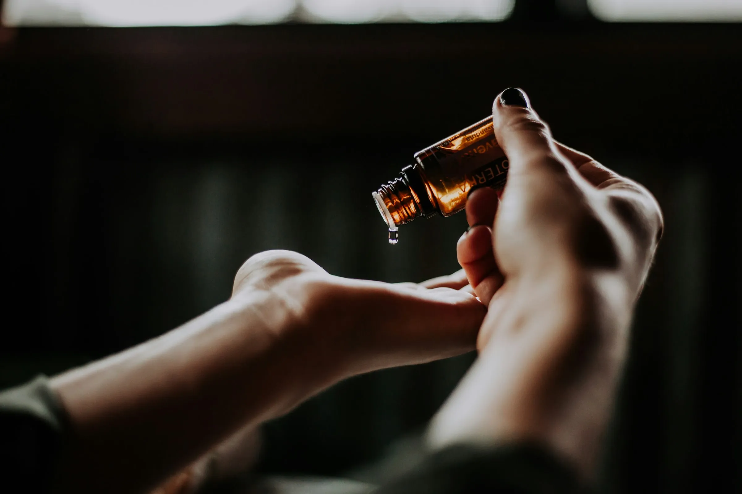 A hand tipping an amber glass bottle to pour a drop of oil into an open palm, in a warm, low-lit room