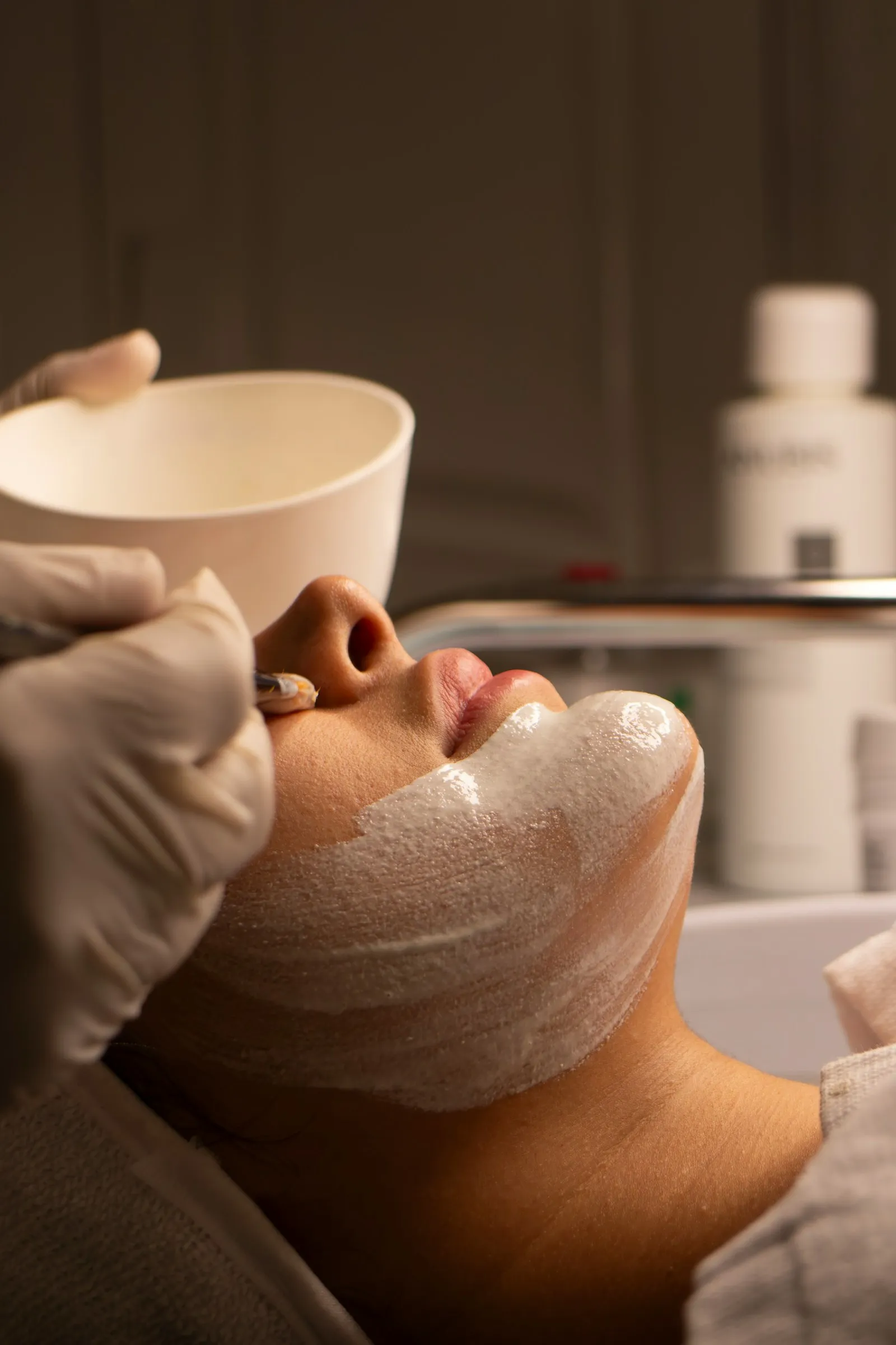 A gloved hand brushing a pale mask along a client's jaw during a facial, the mixing bowl held just above