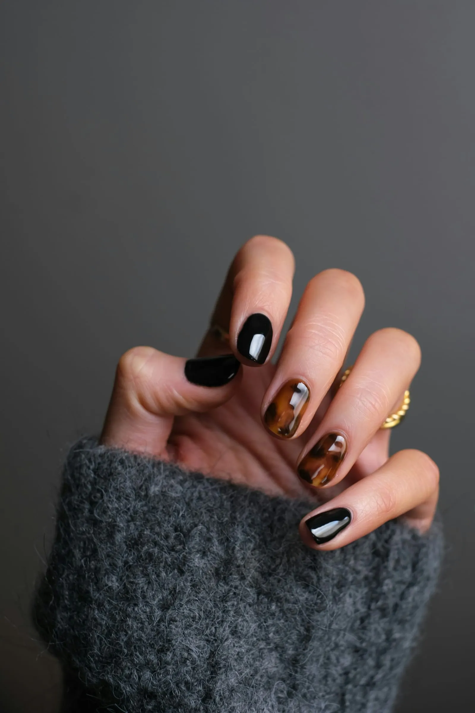 A hand resting against a grey wool sleeve, nails painted in glossy black and tortoiseshell, with a gold ring